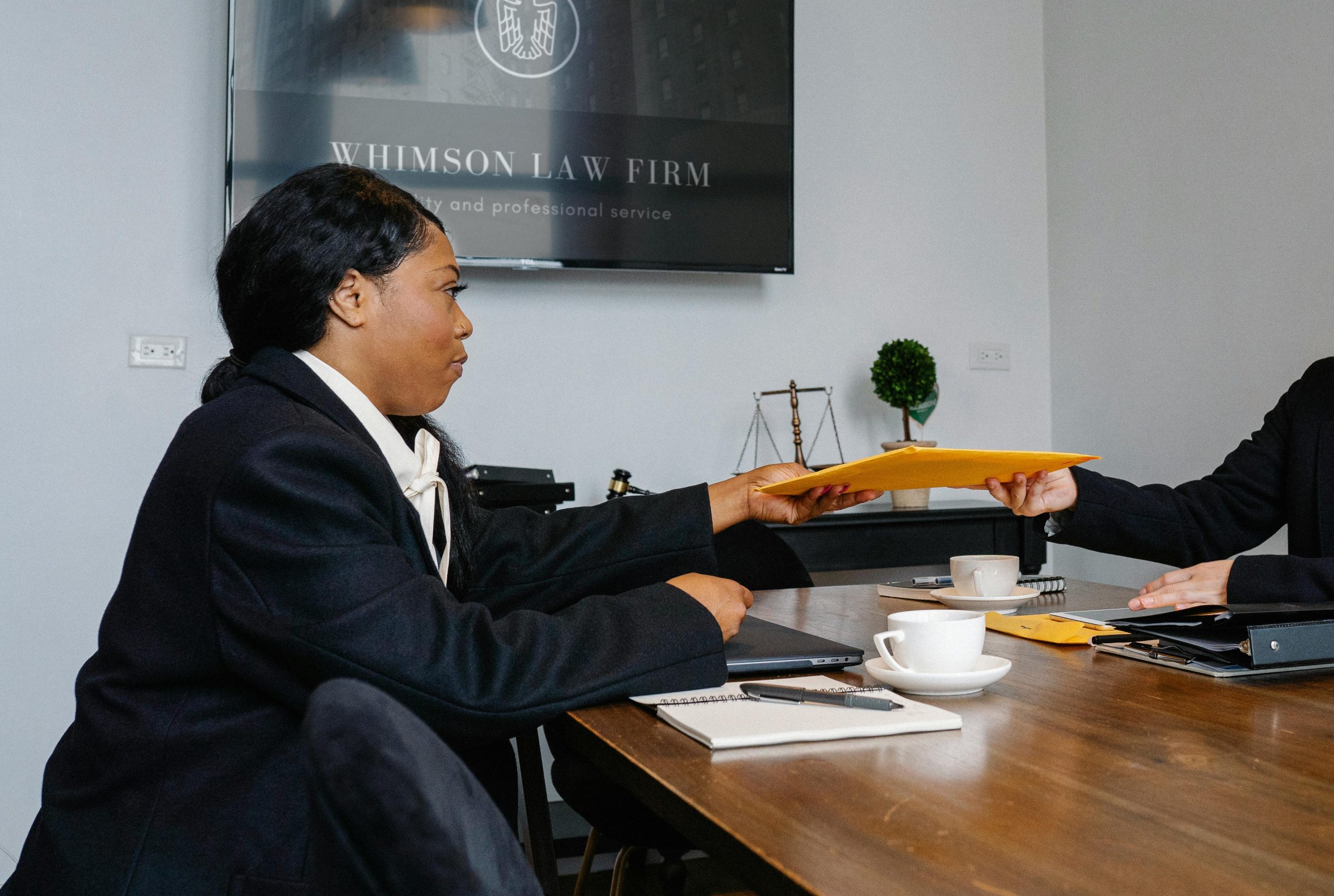 Side view of African American woman in formal wear sitting at table with documents and laptop while surrendering documents to colleague