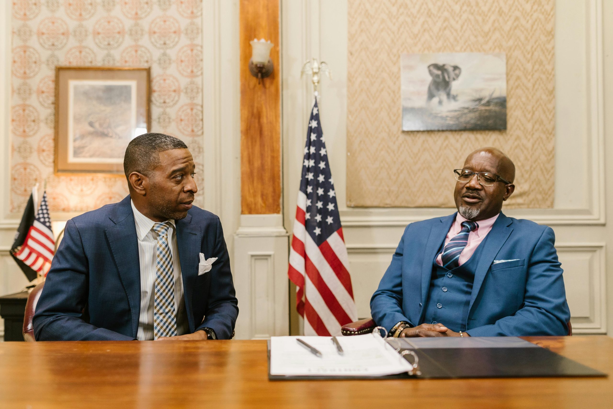 Two businessmen in suits having a discussion at a wooden table with flags in the background.