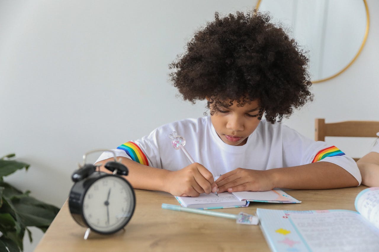 Concentrated African American child writing in notebook while studying at desk with alarm clock at home